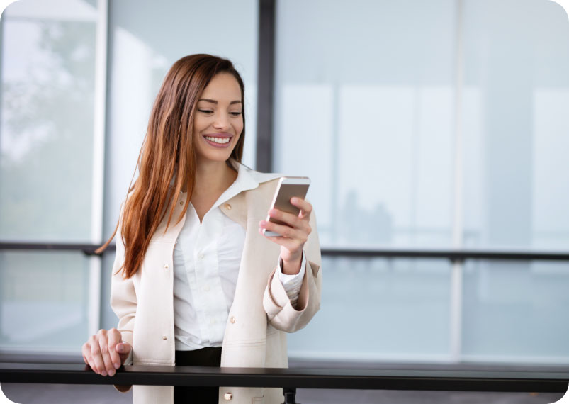 Smiling woman in business casual clothing standing on an office balcony staring at her smartphone