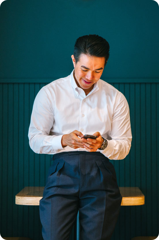 Man in a white shirt and blue pants leans against a desk and a teal wall while texting a colleague