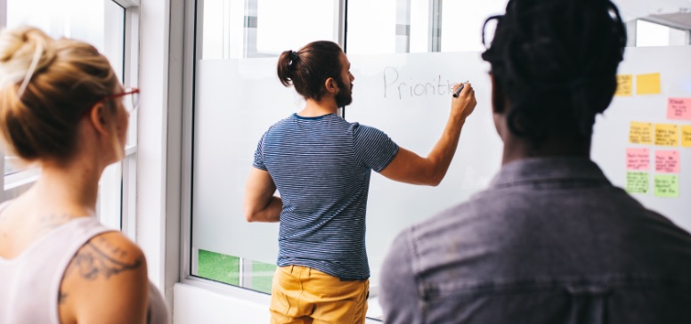 blond women and dark haired man watch a colleague in casual clothes and a man bun draw on a whiteboard