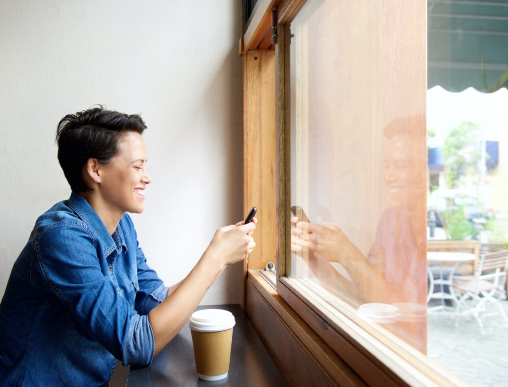 short-haired women happily looking at her phone in front of a window in a cafe