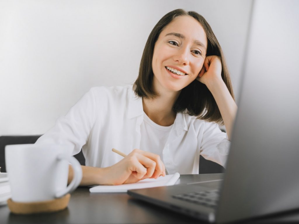 smiling brunette women taking notes in front of a laptop